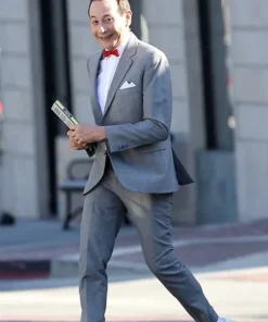 Pee Wee Herman smiling in his signature grey pee wee herman outfit with a red bow tie and white shoes while holding a booklet.