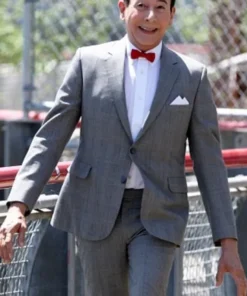 Pee Wee Herman walking outdoors in his classic grey pee wee herman suit with a red bow tie and white shirt.