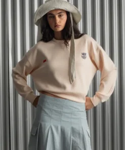 Woman wearing a peach Barça Essentials Crew Sweatshirt with FC Barcelona crest, styled with a light blue skirt and wide-brim hat