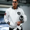 Tom Brady wearing a white Raiders varsity jacket with the team's shield logo patch, walking and holding a water bottle.
