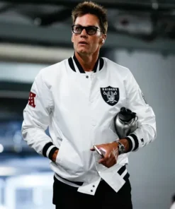 Tom Brady wearing a white Raiders varsity jacket with the team's shield logo patch, walking and holding a water bottle.