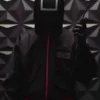 Image of a person wearing the Squid Game black guard suit holding a walkie-talkie, featuring the distinctive black square mask and the hooded black uniform with pink accents. This is a great representation of the black square guard.