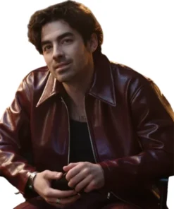 Joe Jonas sitting and smiling faintly while wearing a deep reddish-brown leather jacket with a zip front and collar, over a black tank top. He has a silver ring and a bracelet on, and his hands are clasped. A small chest tattoo is visible.