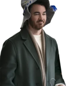 Kevin Jonas sitting indoors and looking contemplative, wearing a dark green coat unzipped over a collared shirt, with his hands clasped in front of him.