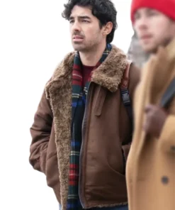 Joe Jonas in a rustic brown shearling-lined suede leather bomber jacket with a furry collar, a dark red sweater underneath, and a plaid green, blue, and red scarf. He is looking off to the side, standing next to a blurred figure wearing a red beanie and a tan coat.