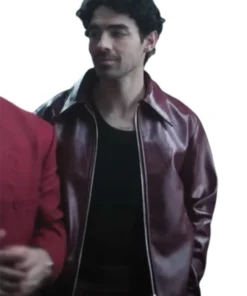 Joe Jonas in a glossy, zippered maroon leather jacket with a wide collar, worn over a black t-shirt. He is looking off to the side, and a person in a red blazer is partially visible on the left.