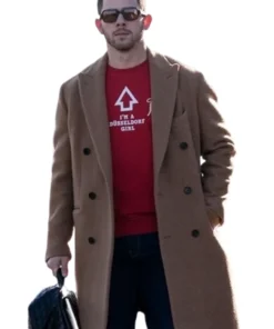 Nick Jonas sitting down, wearing a camel-colored coat over a dark green button-down shirt, dark pants, and a bright red knit beanie. He is looking off to the side, with his arms crossed and some blurred greenery in the foreground.