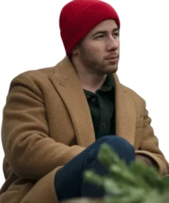 Nick Jonas standing in a long, camel-colored double-breasted coat and dark jeans. He wears dark sunglasses and a red sweatshirt underneath that reads "I'M A DÜSSELDORF GIRL" with an arrow pointing up. He is holding a black bag in his left hand.