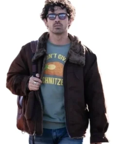 Joe Jonas, wearing sunglasses and a dark brown bomber jacket with a shearling collar, carries a brown leather backpack over his shoulder. He's wearing a light green graphic t-shirt that says "I DON'T GIVE A SCHNITZE" and blue jeans.
