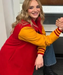 Alana Higman is sitting down and smiling, showing her long red coat with yellow varsity-style sleeves, wide-leg blue jeans, and black ankle boots. The coat's cuffs have red and yellow stripes.