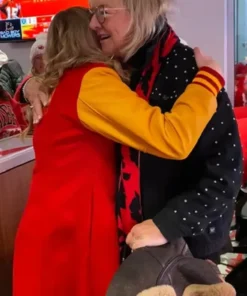Alana Higman, wearing a red coat with yellow sleeves, is embracing an older blonde woman who is wearing glasses, a black jacket with white dots, and a red and black patterned scarf.