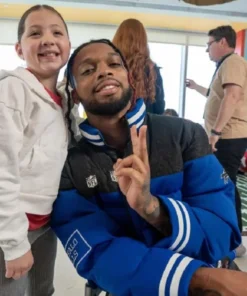 NFL player Damar Hamlin smiles while posing with a young girl. He is wearing a blue and black puffer jacket with a ribbed collar and an NFL shield logo, and he is holding up a peace sign.