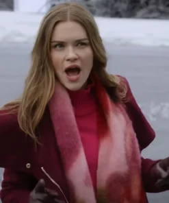 Close-up of Holland Roden looking surprised or excited, with her mouth open. She is outdoors in a winter setting, wearing a dark red or burgundy coat, a bright pink turtleneck, a fuzzy pink and red scarf, and dark gloves.