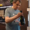 Close-up of Rowan Atkinson as Trevor Bingley in the comedy series Man vs Baby 2025, wearing a light blue/grey, round-neck Fish Printed T-Shirt while holding a bottle of sparkling wine. The T-Shirt features a horizontal print of stylized fish.