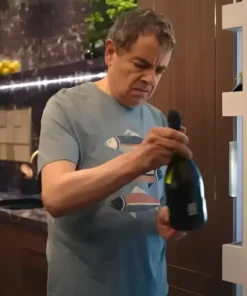 Close-up of Rowan Atkinson as Trevor Bingley in the comedy series Man vs Baby 2025, wearing a light blue/grey, round-neck Fish Printed T-Shirt while holding a bottle of sparkling wine. The T-Shirt features a horizontal print of stylized fish.
