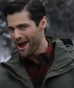 Close-up of actor Matthew Daddario looking surprised or excited, with his mouth open. He is wearing an olive green hooded work jacket with a small brand logo on the chest, layered over a red plaid shirt.