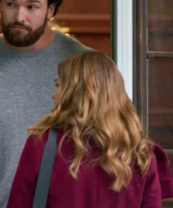 Holland Roden, with long wavy strawberry-blonde hair, is seen from the back wearing a burgundy or deep red coat and a shoulder bag. A tall man with a full beard and a gray sweater is standing behind her, looking over her shoulder.