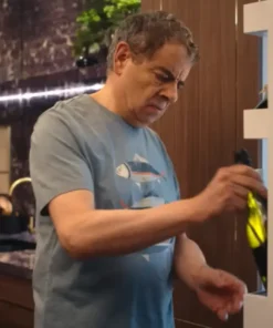 Mid-shot of Rowan Atkinson looking concerned while pulling a bottle from a wine rack. He is wearing a light blue/grey, crew-neck Fish Printed T-Shirt which features a graphic of stylized fish. This image highlights the casual Rowan Atkinson T-Shirt outfit.