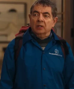 Close-up of Rowan Atkinson from "Man vs Baby," wearing the distinctive Housesitters Deluxe Blue Jacket with the logo visible. He is wearing a backpack and a patterned sweater underneath the Rowan Atkinson blue jacket for men.