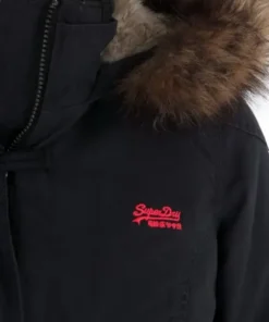 A close-up shot of the shoulder and collar of the black Superdry Study Duffle Jacket. It clearly shows the zipper detail, the textured brown Vegan fur hood trim, and the small, bright red embroidered Superdry logo on the chest.