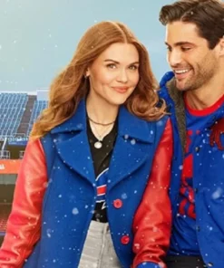 Holland Roden and Matthew Daddario pose together in front of a snowy stadium background. Roden, with long red hair, smiles while wearing a striking royal blue coat with red leather sleeves over a black Buffalo Bills t-shirt. Daddario smiles beside her, also wearing a blue and red jacket.
