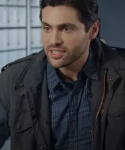 Close-up of actor Matthew Daddario looking sternly off-camera. He has dark hair and a slight beard, and is wearing a dark, possibly black or charcoal, field jacket over a blue plaid button-down shirt.