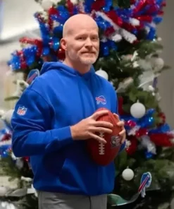 Sean McDermott, the bald-headed coach of the Buffalo Bills, stands in front of a Christmas tree decorated with red, white, and blue ornaments and lights. He is wearing a bright blue Buffalo Bills hooded sweatshirt with the team logo on the chest and an NFL shield patch on the sleeve, holding a football with both hands.