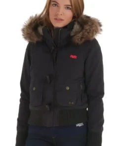 A young woman posing in a short, black Superdry Study Duffle Jacket. The jacket is a puffer style with a ribbed waistband and cuffs, large front flap pockets with toggle closures, and a hood trimmed with thick brown Vegan fur. The red embroidered Superdry logo is visible on the chest.