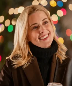 A close-up portrait of Alexandra Breckenridge smiling, wearing a thick black turtleneck sweater and a chocolate brown wool coat against a backdrop of blurred holiday lights.