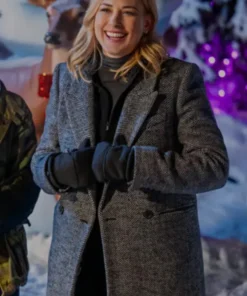 Smiling woman wearing a grey herringbone double-breasted winter coat with black gloves and a high-neck top in a snowy holiday setting.