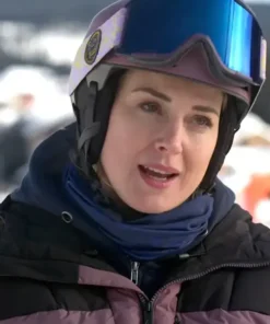 Woman wearing a ski helmet and goggles with a black and mauve puffer jacket and blue neck warmer outdoors.