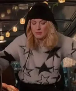 Blonde woman wearing a white star sweater and black beanie in a festive outdoor setting with string lights.