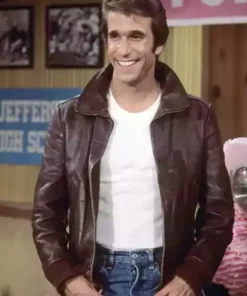 Henry Winkler as Fonzie wearing his signature brown leather jacket over a white t-shirt and blue jeans, standing and smiling on the Happy Days set.