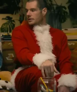 Man sitting indoors wearing a red Santa-style coat with white fur trim in a holiday movie scene.