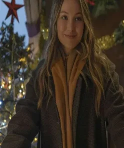 Olivia Holt Jingle Bell Heist long coat in a close-up shot. Sophia wears the Brown Long Coat and a scarf, smiling with Christmas lights behind her.