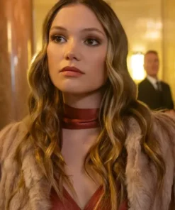 A woman with long wavy hair wears a brown fur coat over a red dress in a warmly lit indoor setting.