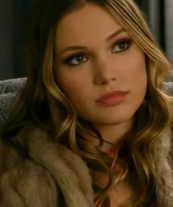 A woman with long wavy hair in a brown fur coat looks ahead while seated indoors.