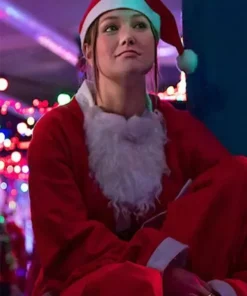 Olivia Holt in a red Santa coat and hat sits with her arms crossed in a brightly lit festive space.